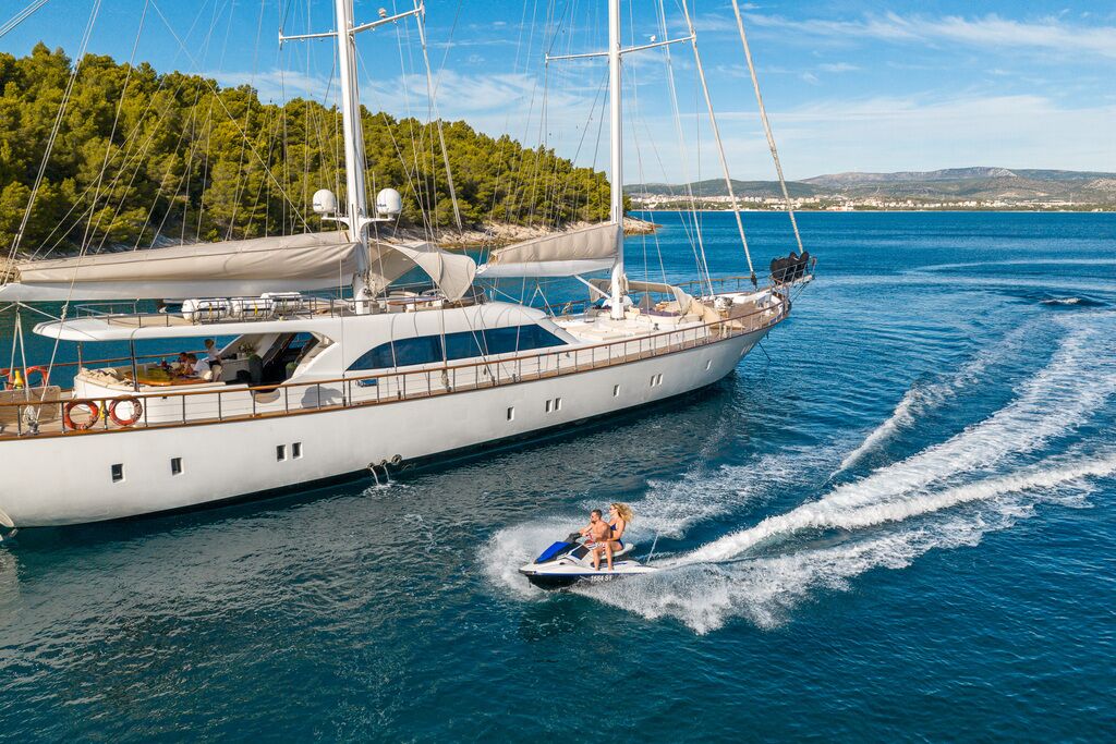 A luxury yacht is anchored near a forested shoreline as two people ride a jet ski on the clear blue water, leaving a trail of white foam behind them under a sunny sky.