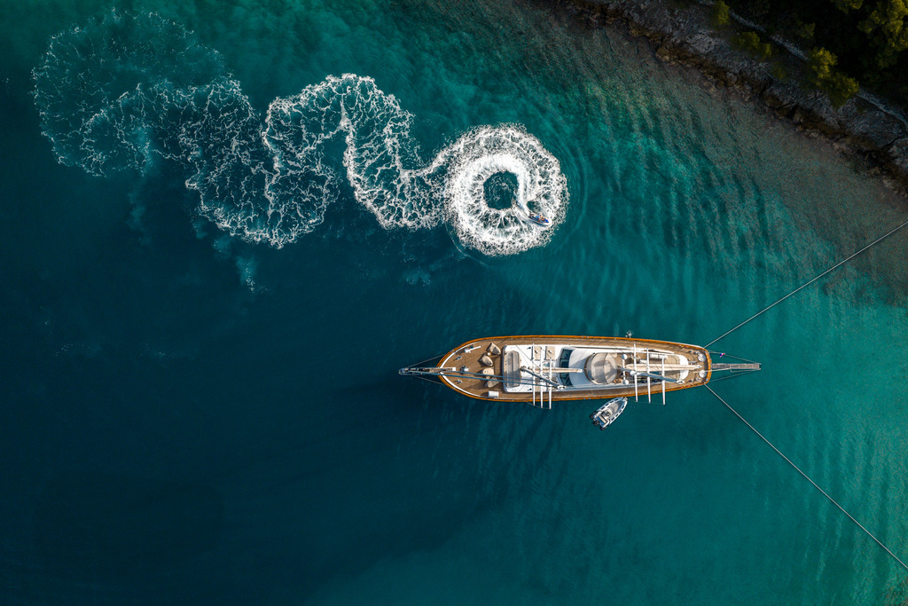 Aerial view of a large yacht anchored in turquoise water near a forested shoreline, with a jet ski creating swirling white waves beside the boat.