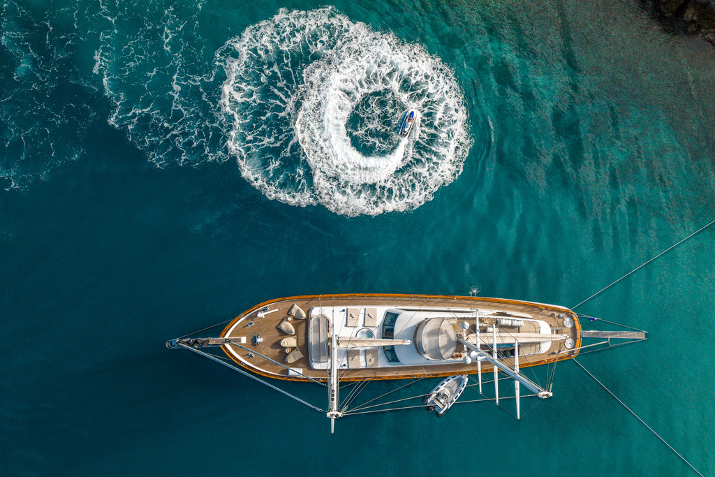 Aerial view of a yacht anchored in clear blue water, with a person on a jet ski making a circular pattern of white waves nearby.