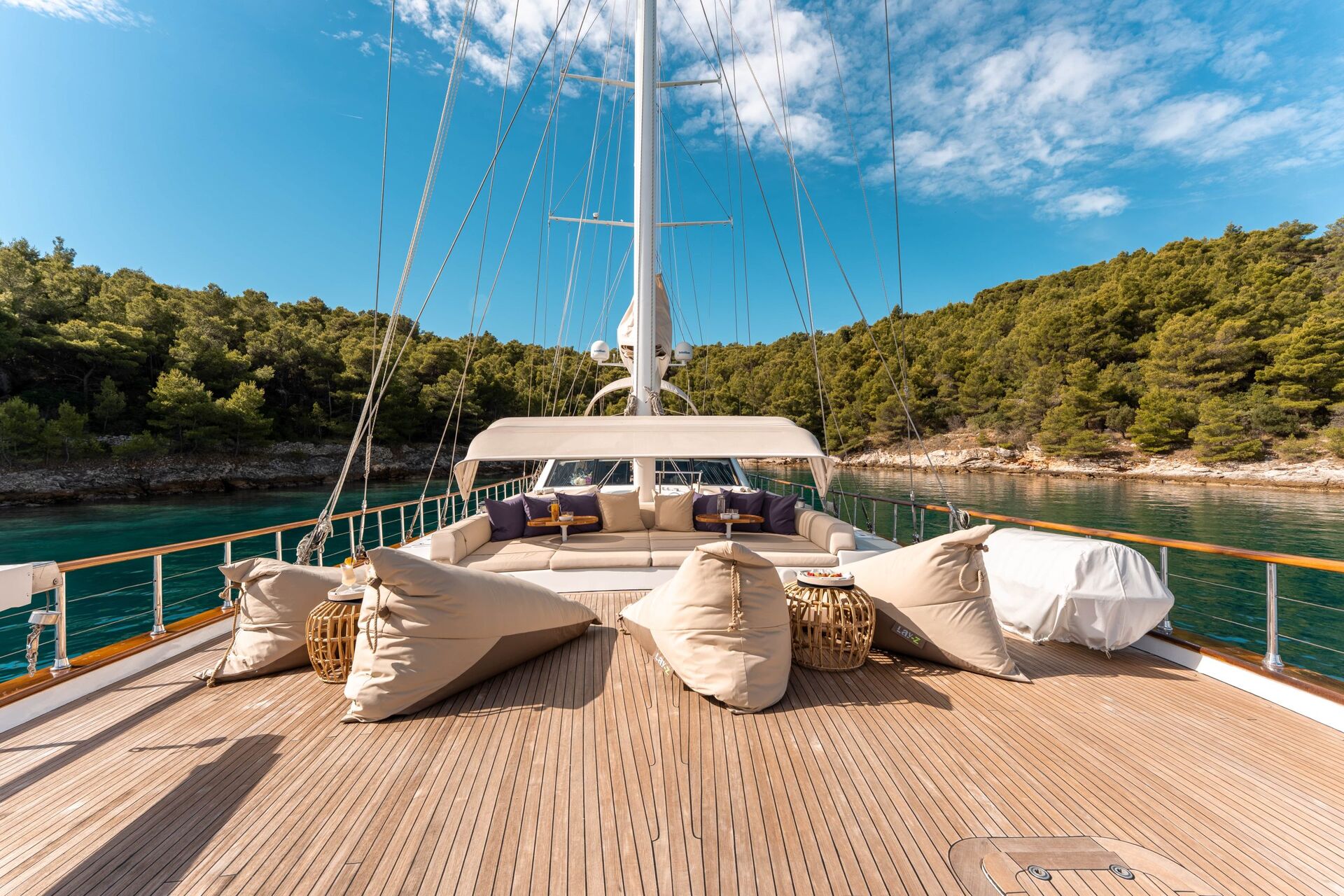 View from the deck of a yacht featuring beige bean bag chairs, a shaded seating area, and tables, with calm turquoise water and a forested shoreline in the background under a blue sky.
