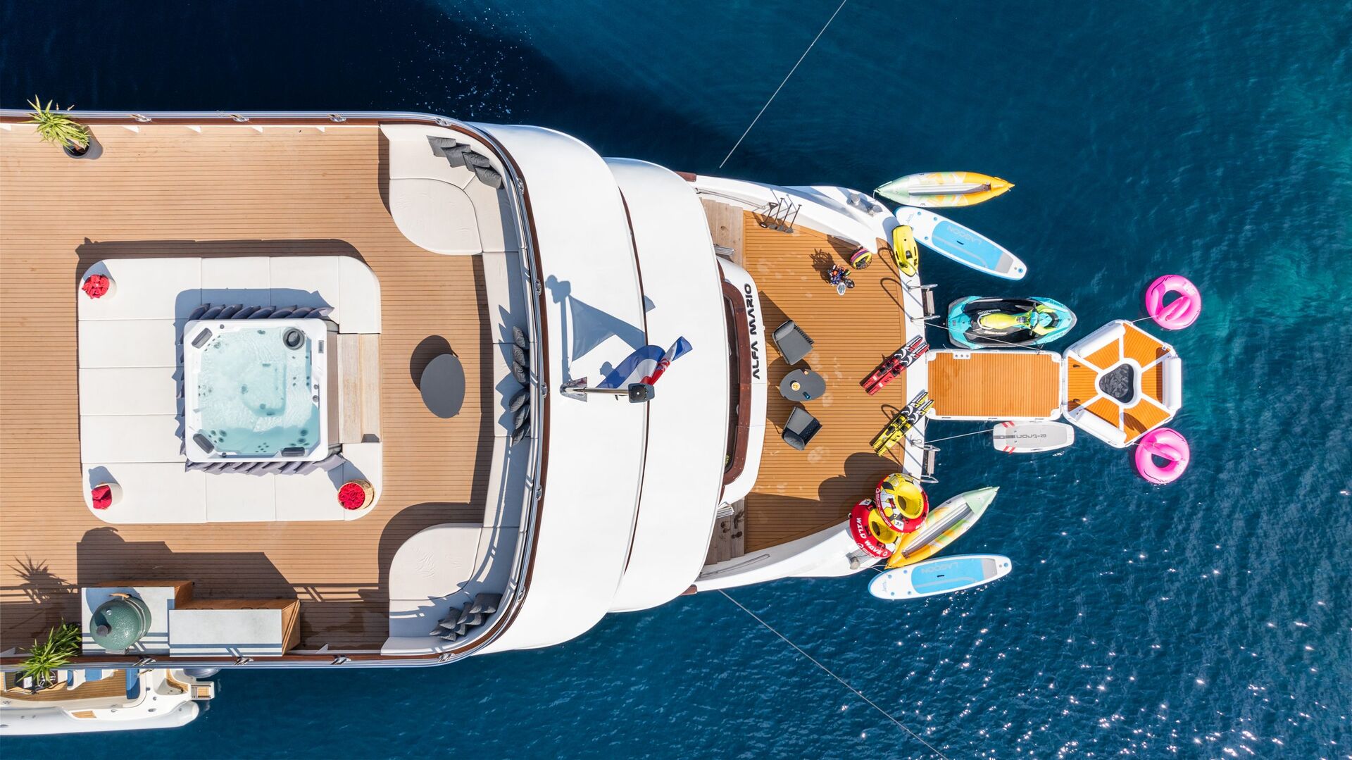 Aerial view of a luxury yacht on blue water, featuring a hot tub, sun loungers, water toys, paddleboards, and colorful inflatables on the deck and in the water.