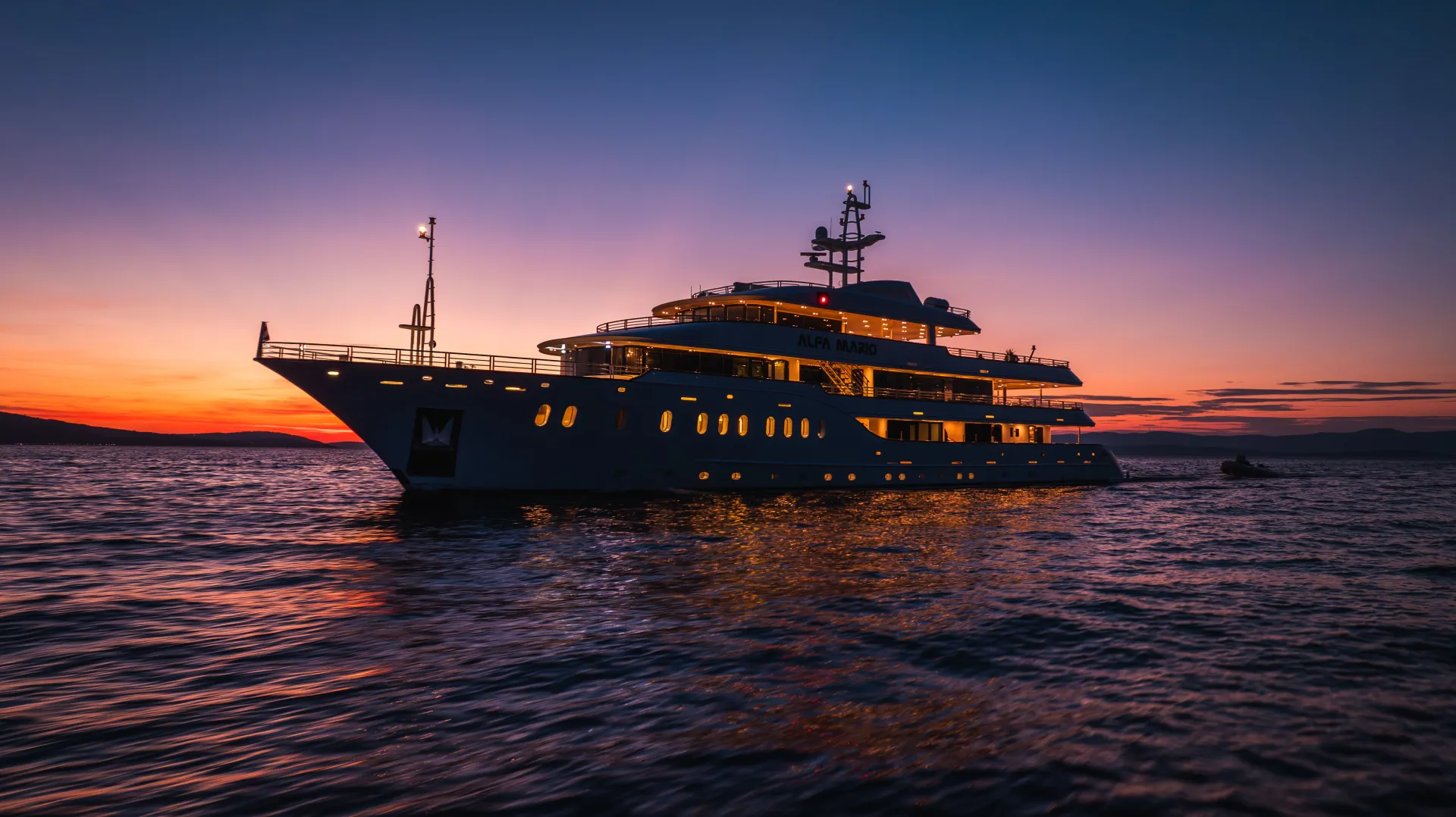 A large luxury yacht is illuminated and anchored on calm water during a colorful sunset, with the sky glowing in shades of orange, pink, and purple.