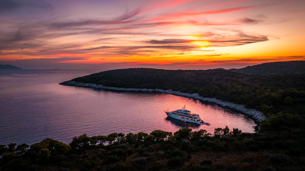 A yacht is anchored near a forested coastline at sunset, with vibrant orange and purple colors in the sky reflecting on calm water. Hills surround the bay, and the scene is serene and peaceful.