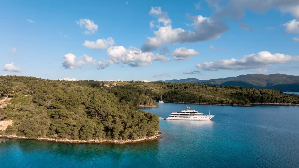 A luxury yacht is anchored in a calm, blue bay surrounded by lush green forest and hills under a partly cloudy sky. The water is clear, and the shoreline is rocky with dense trees.
