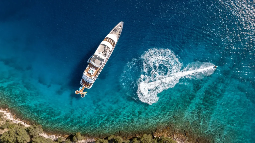 Aerial view of a large yacht anchored in clear blue water near a coastline, with a jet ski creating a circular wake pattern nearby over the turquoise sea. Dense greenery lines the shore at the bottom of the image.