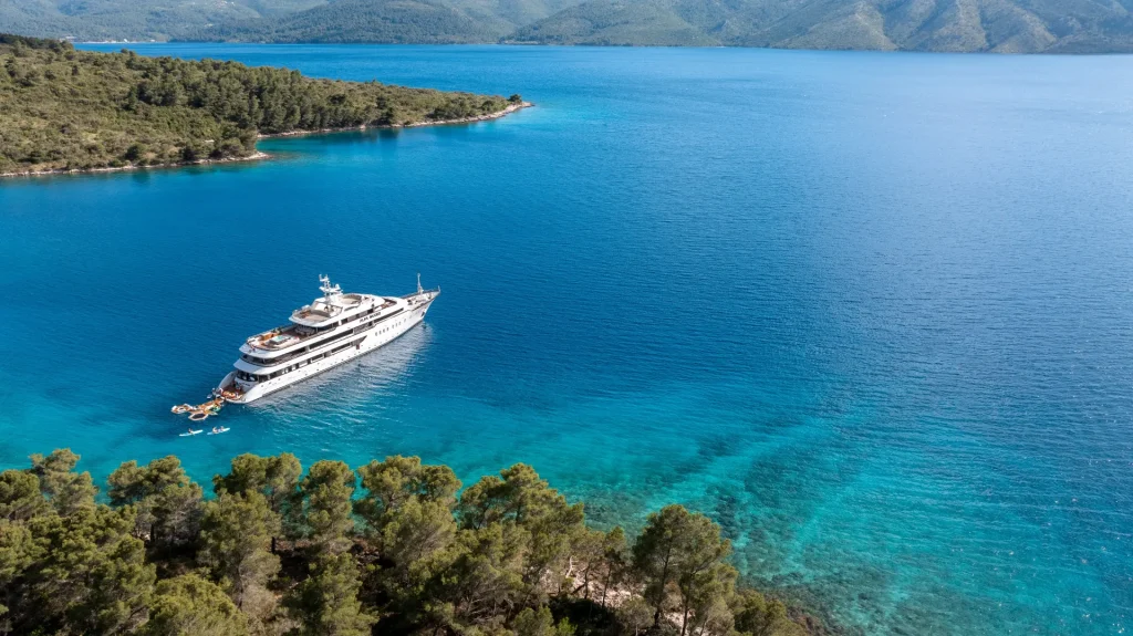 A luxury yacht is anchored in clear blue water near a lush, green coastline, with forested hills and distant mountains in the background under a bright sky.