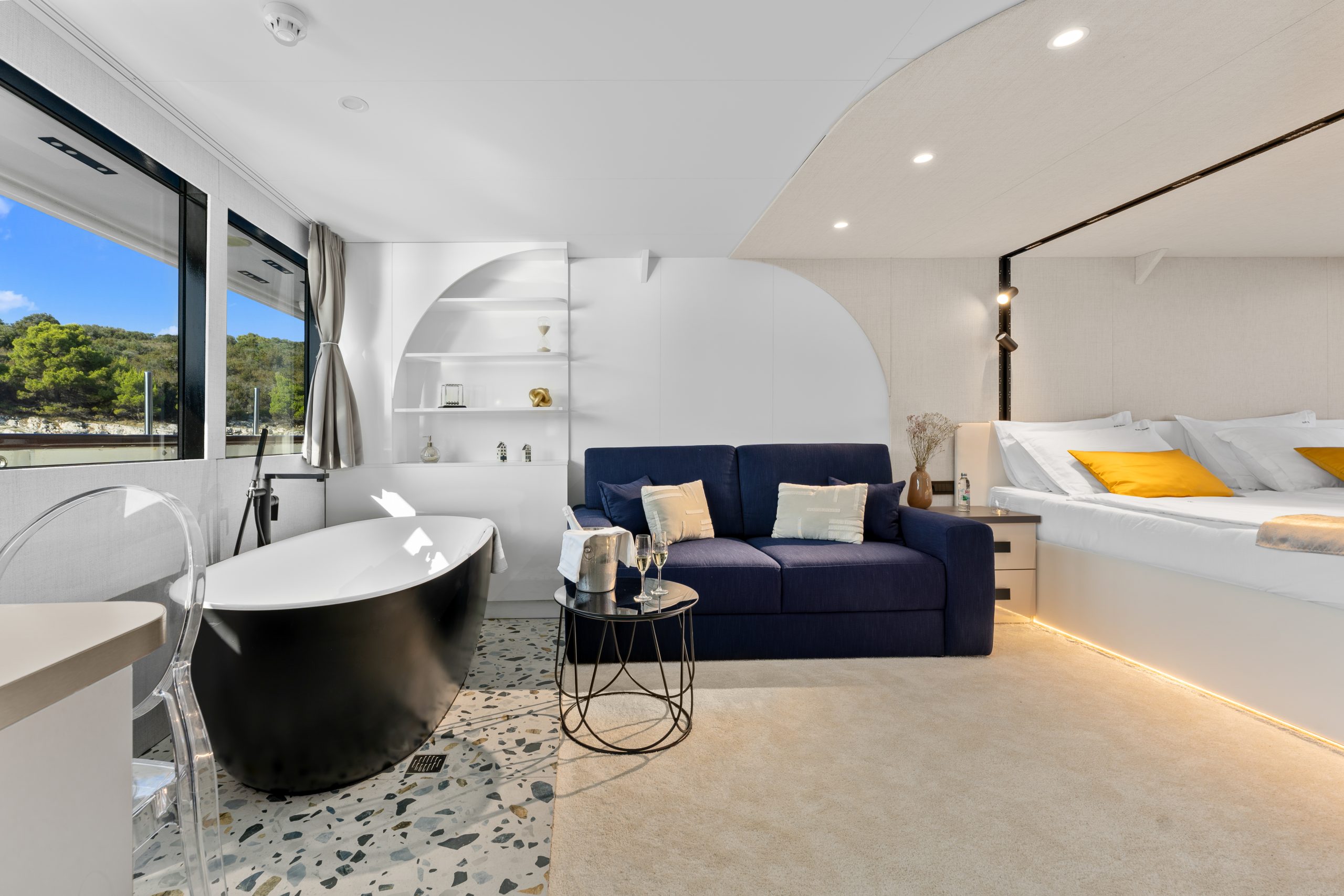 Modern hotel room with a black freestanding bathtub, blue sofa, round glass coffee table, double bed with yellow pillows, built-in shelves, and large window with a view of trees outside.