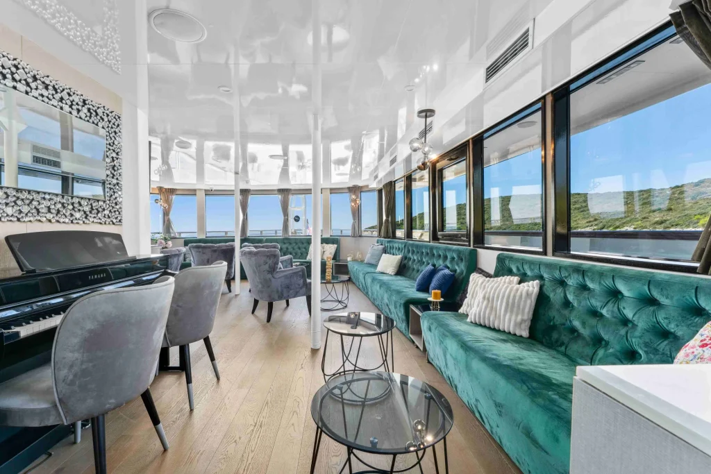 Modern lounge with teal velvet sofa, gray armchairs, a black piano, and large windows offering views of greenery and blue sky. The room has wooden floors, white ceiling, and elegant decor.