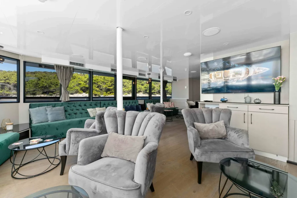 Modern yacht lounge with large windows, plush gray armchairs, green sofa, glass coffee tables, light wood floors, and a flat-screen TV displaying a boat. Sunlight streams in, and greenery is visible outside.