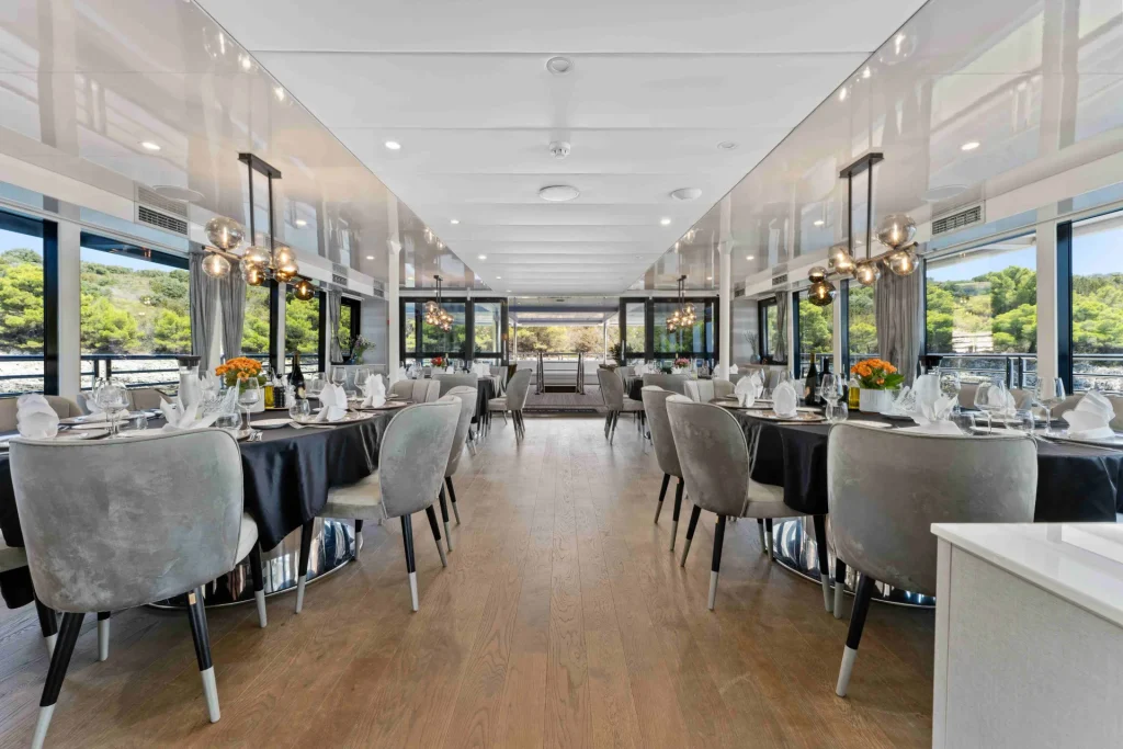 A modern, elegant dining room on a yacht with large windows, round tables set with white linens and flowers, gray velvet chairs, and scenic views of trees and water outside.