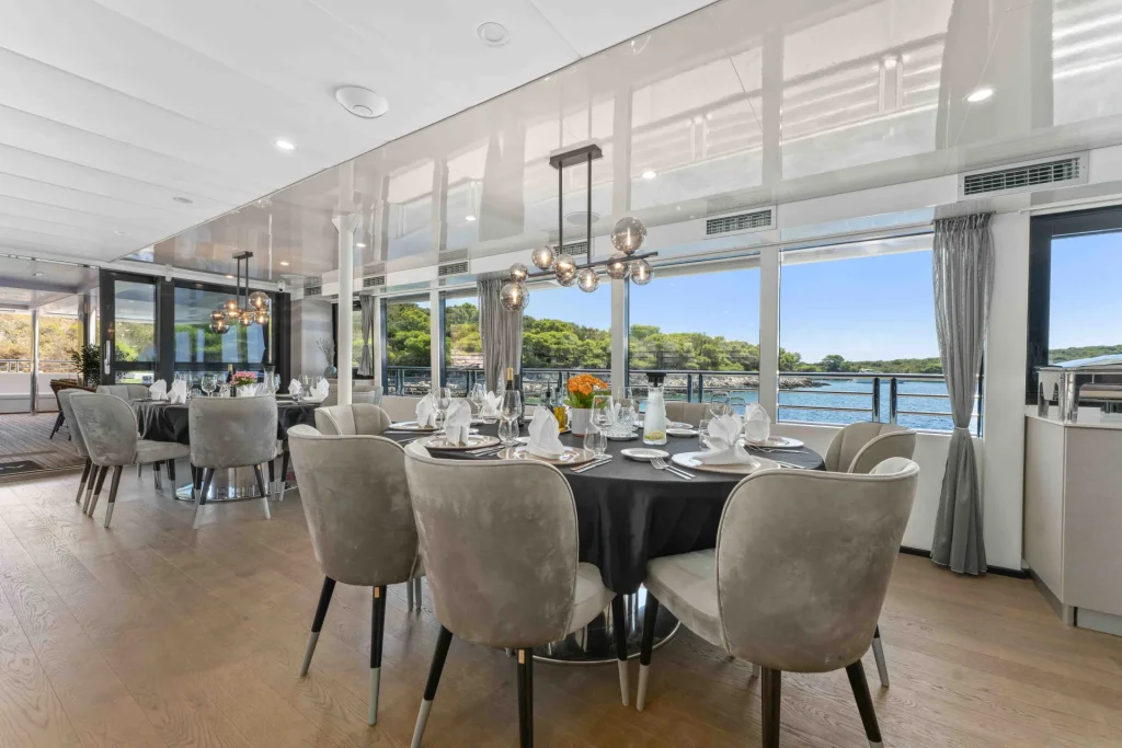 A modern dining area with round tables set for a meal, elegant gray chairs, large windows showing a scenic waterfront view, and contemporary lighting fixtures hanging from the ceiling.
