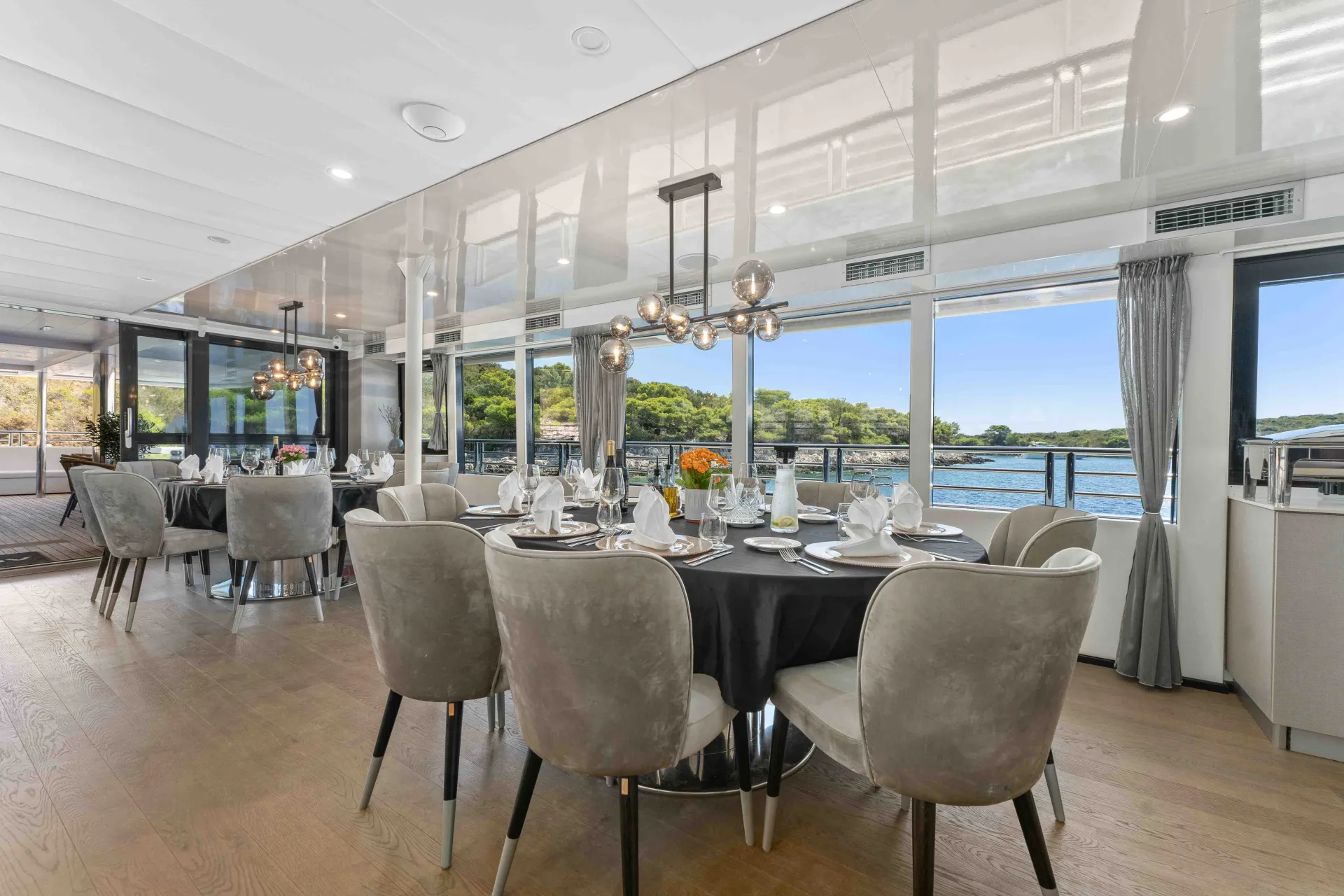 A modern dining area with round tables set for a meal, elegant gray chairs, large windows showing a scenic waterfront view, and contemporary lighting fixtures hanging from the ceiling.