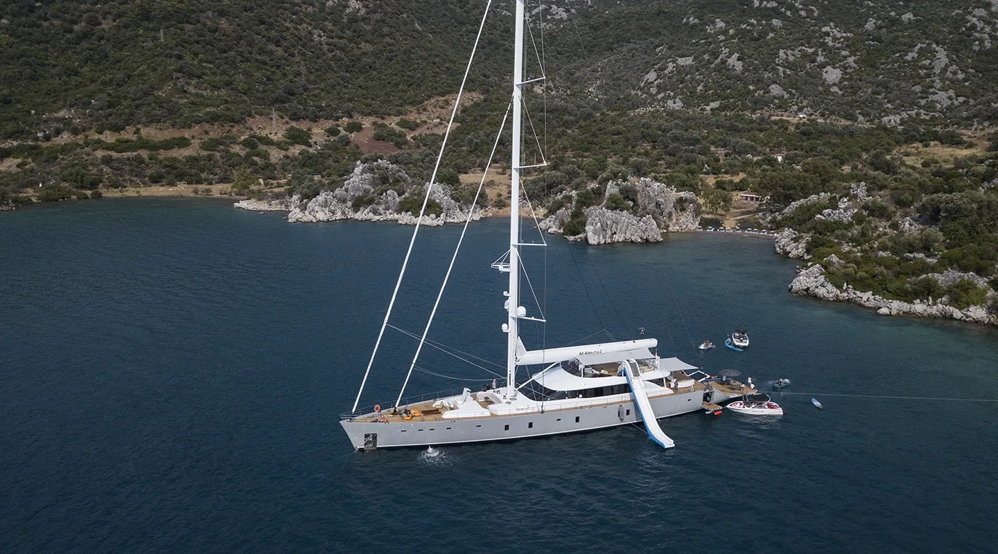 A white sailboat with a tall mast is anchored near a rocky, forested shoreline. Several small boats float nearby, and a slide extends from the yacht into the blue water. Hills and greenery are visible in the background.