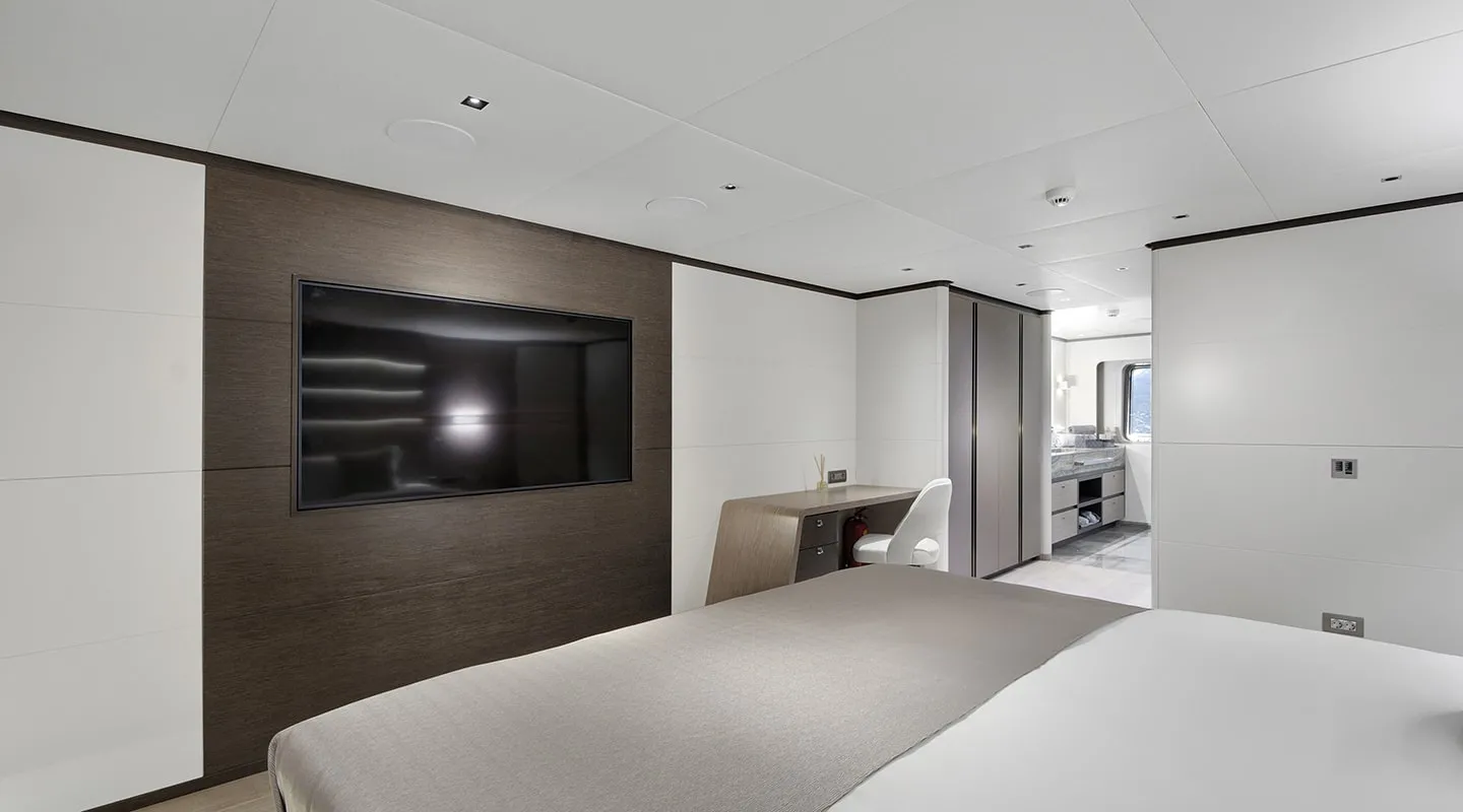 A modern, minimalist bedroom with a large wall-mounted TV, a desk and chair, neutral-colored bedding, and an en suite bathroom visible in the background. The decor features clean lines and muted tones.