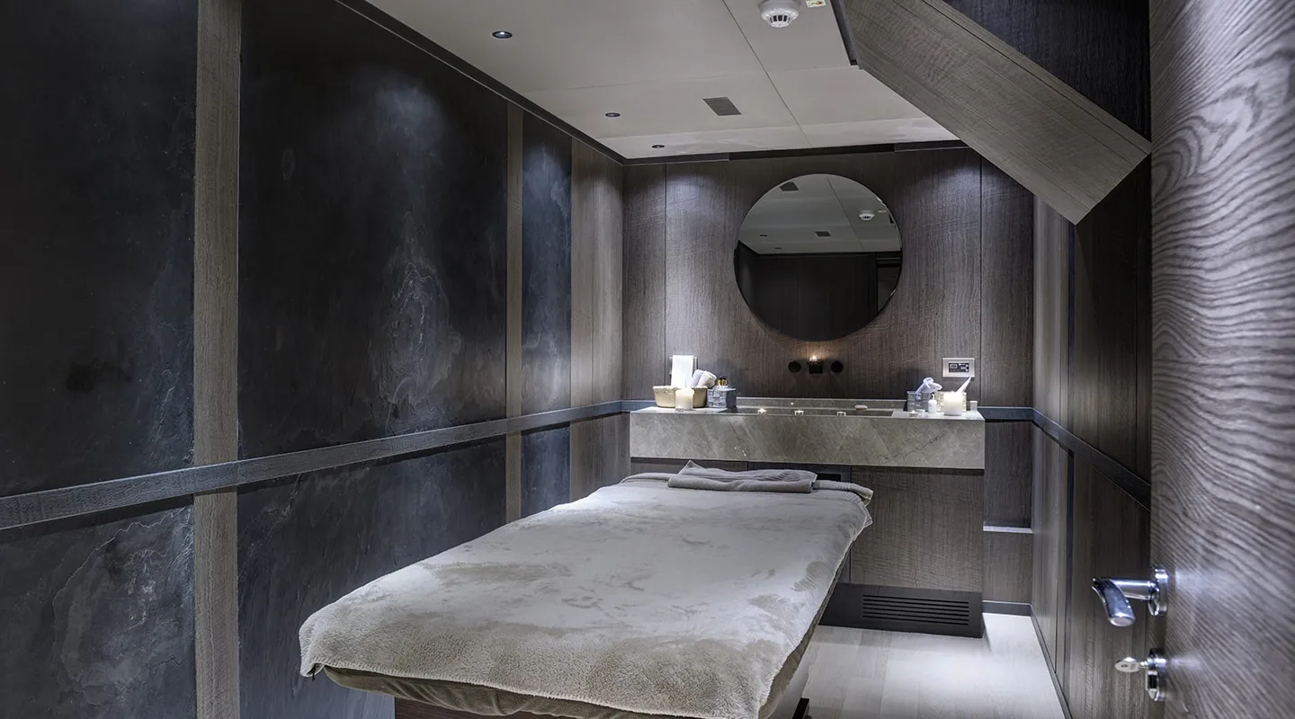 A modern, dimly lit spa room with dark wood walls, a massage table covered in a beige towel, a counter with spa supplies, and a round mirror on the wall above the counter.
