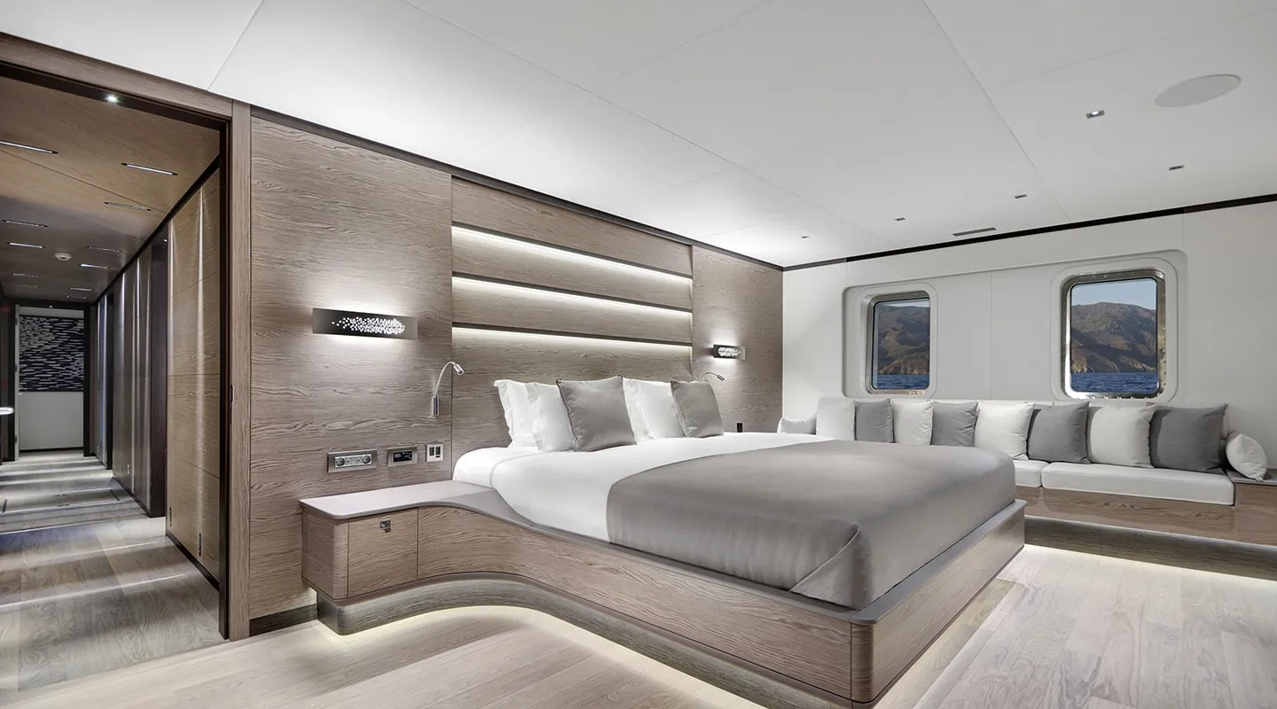 A spacious, modern yacht bedroom with a large bed, neutral tones, built-in lighting, a long cushioned bench under three windows, and minimalist wooden décor, with a scenic view of mountains and water outside.