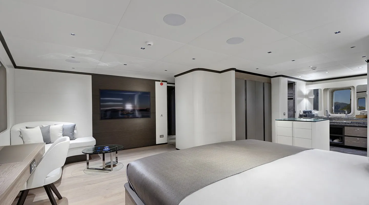 A modern, spacious bedroom suite with a large bed, wall-mounted TV, desk with chair, small sofa with glass table, built-in closets, and windows offering scenic views. Decor is minimalist with neutral tones.