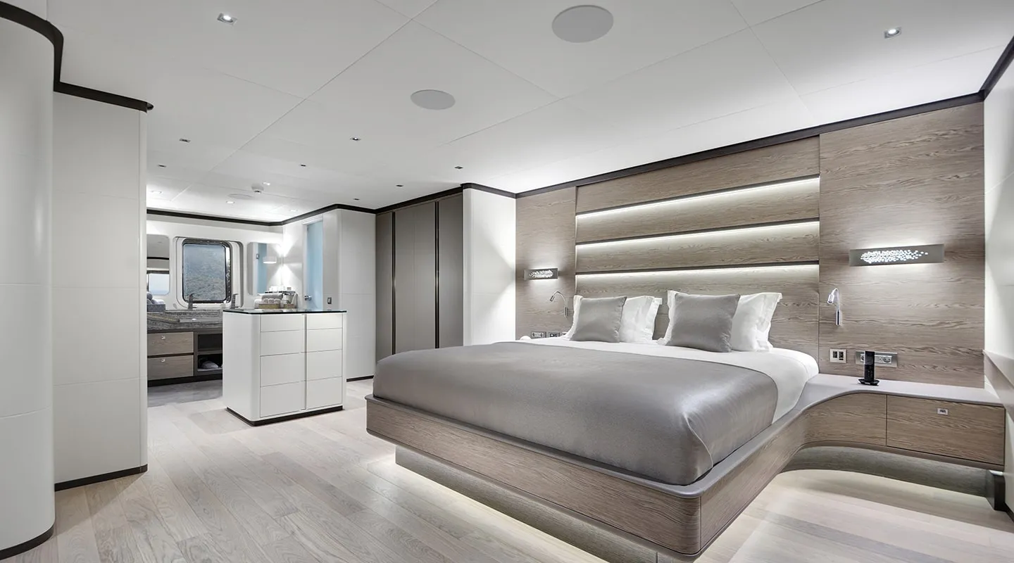 Modern, minimalist bedroom with a large platform bed, neutral colors, built-in wall lighting, sleek cabinetry, and a spacious en suite bathroom visible in the background.