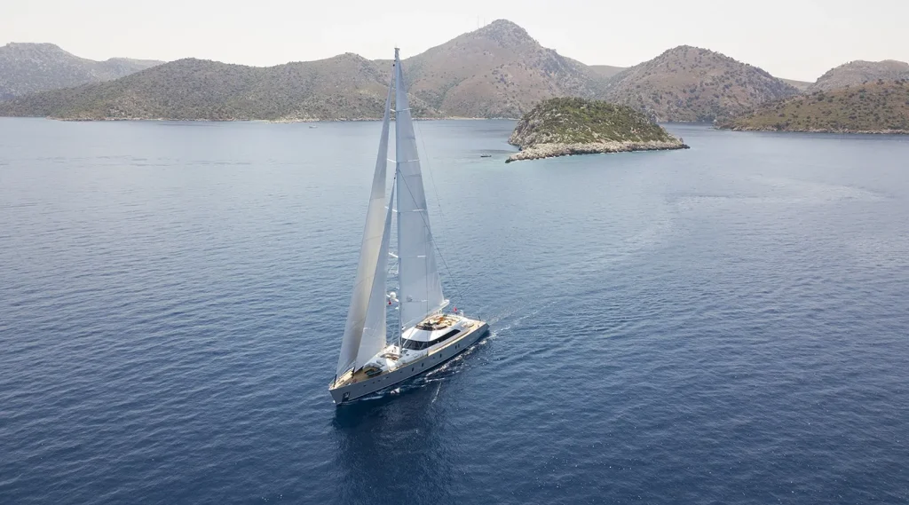 A large sailboat with white sails glides across calm blue water, surrounded by green, hilly islands under a clear sky.