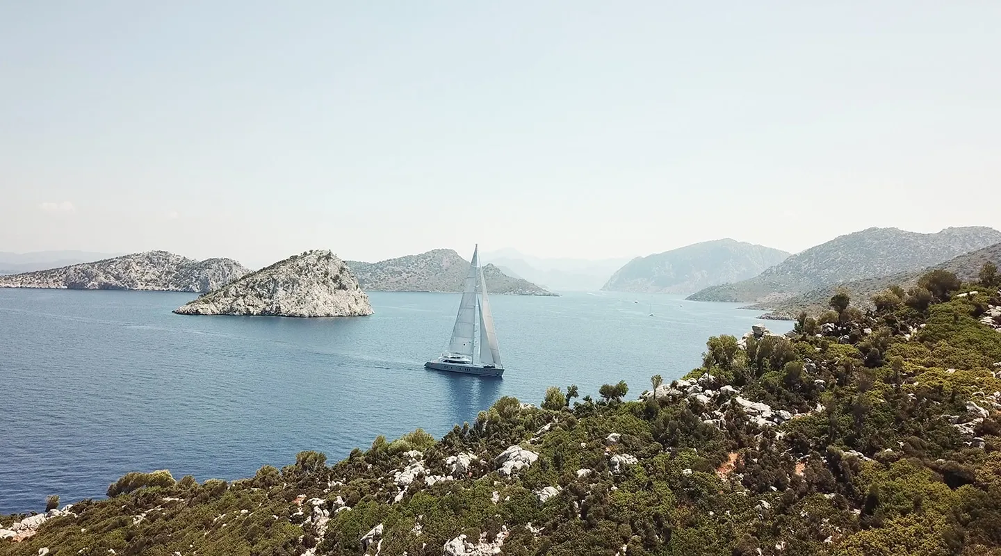 A sailboat glides across calm blue water surrounded by rocky green hills and small islands under a clear sky, creating a peaceful coastal landscape.