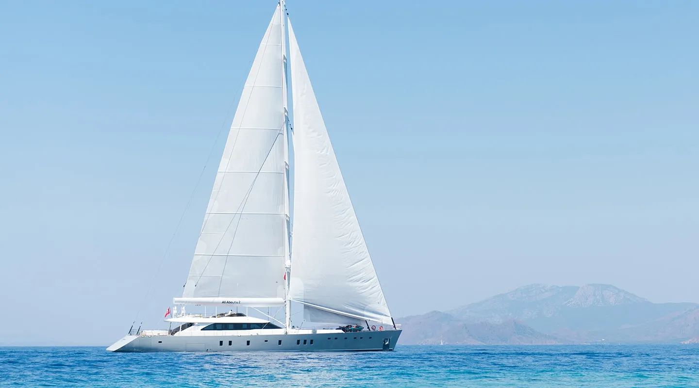 A large white sailboat with fully raised sails glides on calm blue water, with distant mountains visible in the background under a clear sky.