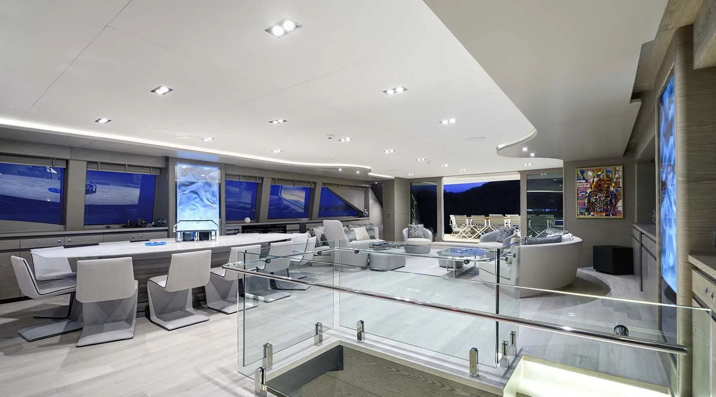 Modern, spacious yacht interior with large windows, sleek white and gray furniture, glass stair railing, lounge and dining areas, and colorful artwork on the wall. Evening light is visible through the windows.