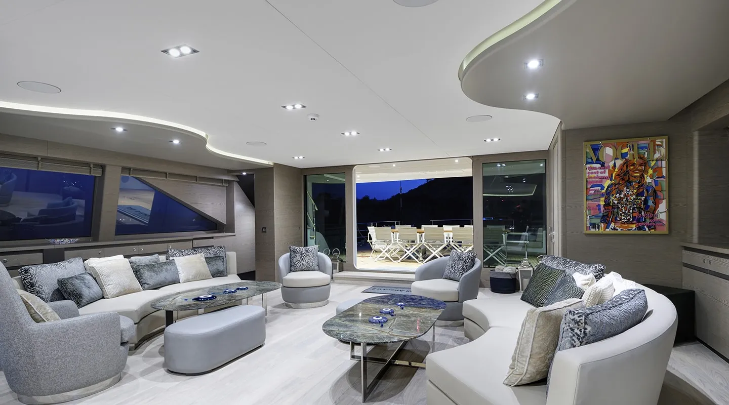 A modern yacht lounge with curved sofas, armchairs, glass tables, decorative cushions, abstract art on the wall, and large windows overlooking an outdoor deck with white chairs. The room is softly lit and elegant.