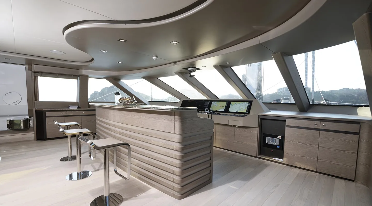 Modern yacht interior with wooden finishes, bar counter with stools, large windows offering scenic views, and multiple navigation screens on the control panel.