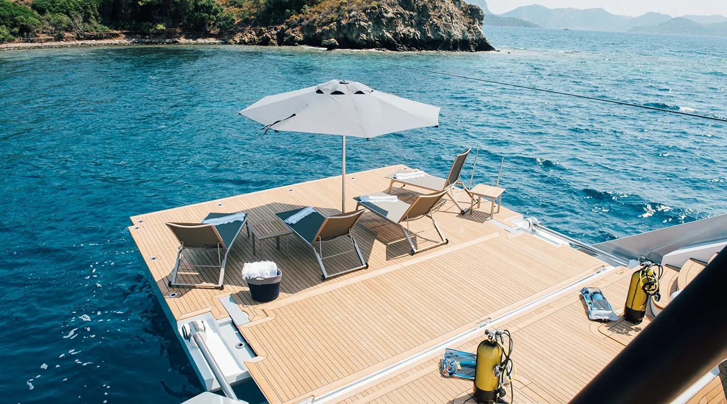 A wooden deck with lounge chairs and an umbrella floats on clear blue water near a rocky coastline, with scuba diving tanks and gear visible in the foreground.
