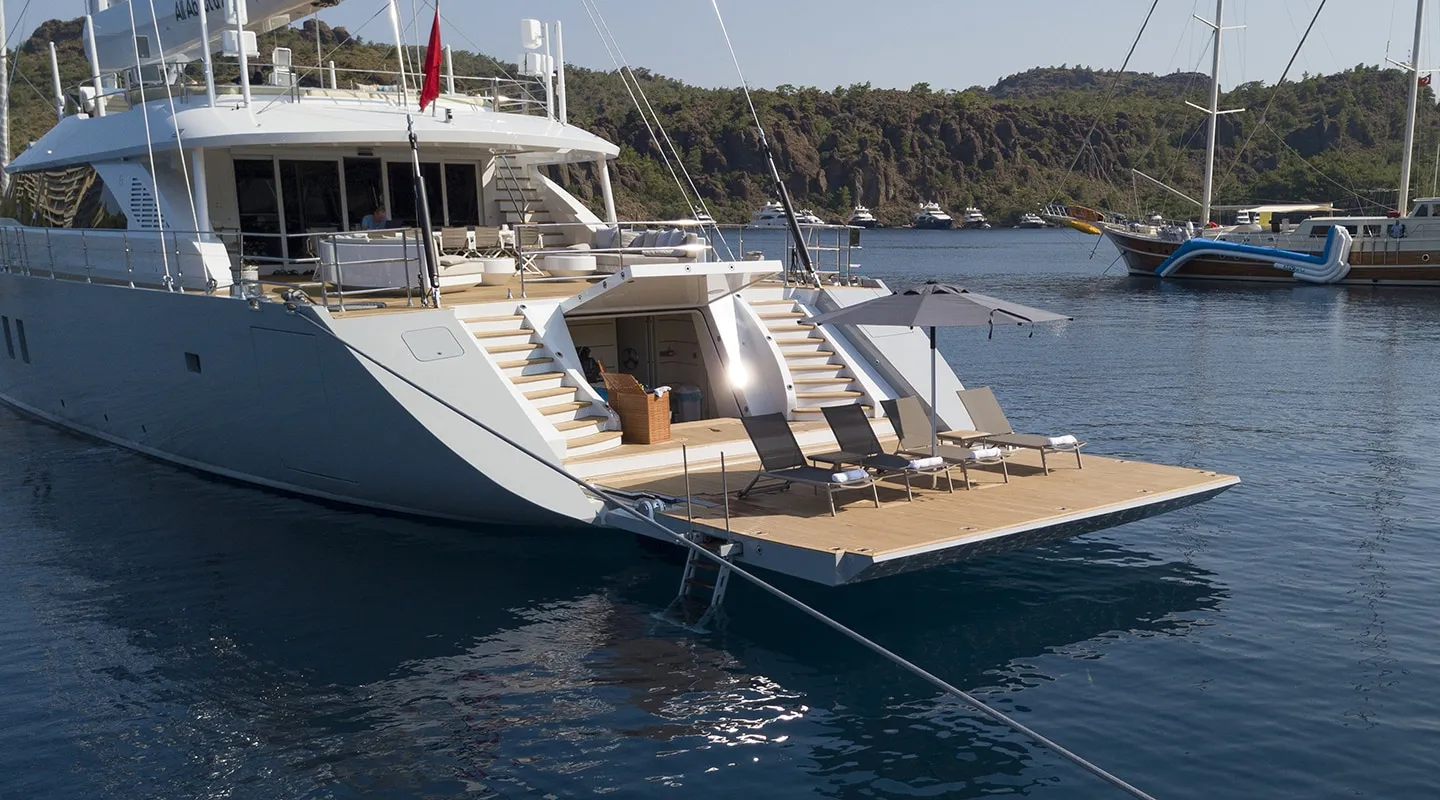 A luxury yacht anchored near the shore features a spacious rear deck with lounge chairs and an umbrella, overlooking calm blue water and a forested coastline.