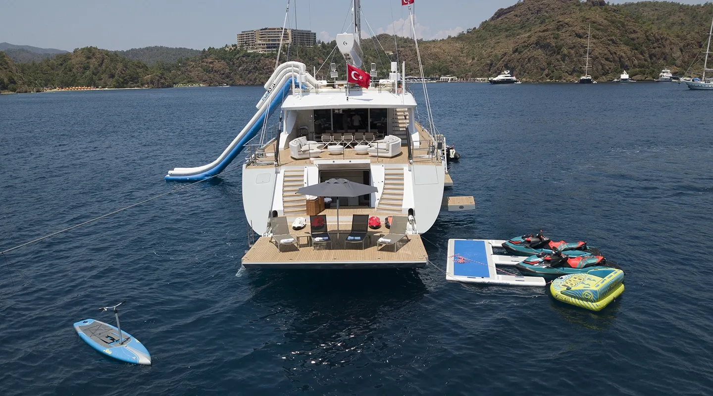 A large yacht anchored in calm blue water, with a waterslide extending from its deck, inflatable toys, jet skis, and a paddleboard nearby. Mountains and other boats are visible in the background.