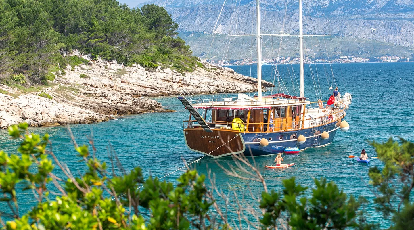 A wooden sailboat named “Altair” is anchored in a turquoise cove near a rocky, tree-covered shoreline; people enjoy swimming and kayaking nearby under a clear sky, with mountains in the distance—perfect for yacht charter enthusiasts.