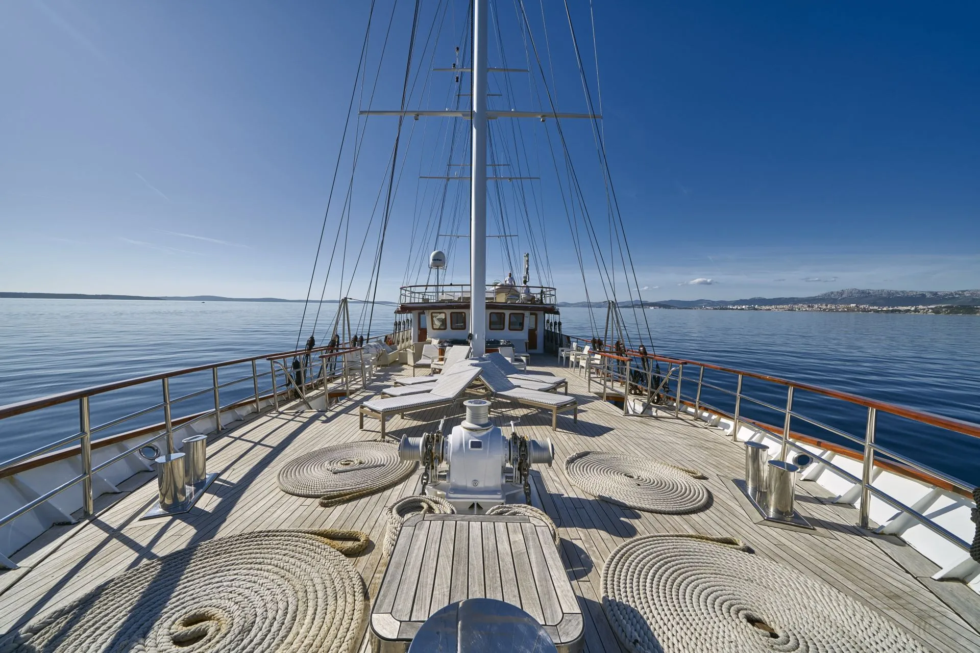 View from the deck of a yacht with neatly coiled ropes, metal railings, and equipment, sailing on calm blue water under a clear sky, with a distant shoreline visible.