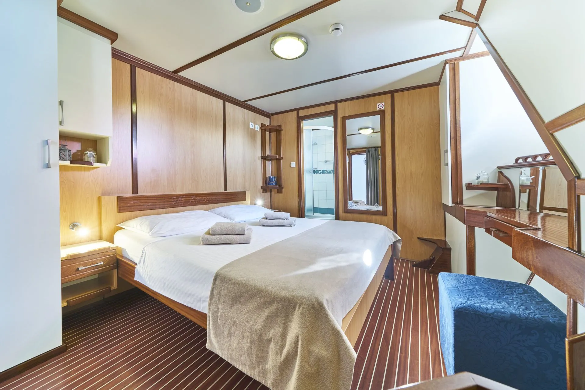 A cozy bedroom with wooden walls and floor features a double bed neatly made with white linens and beige throw, two towel sets, a mirror, a desk with a blue stool, and a staircase in the corner.