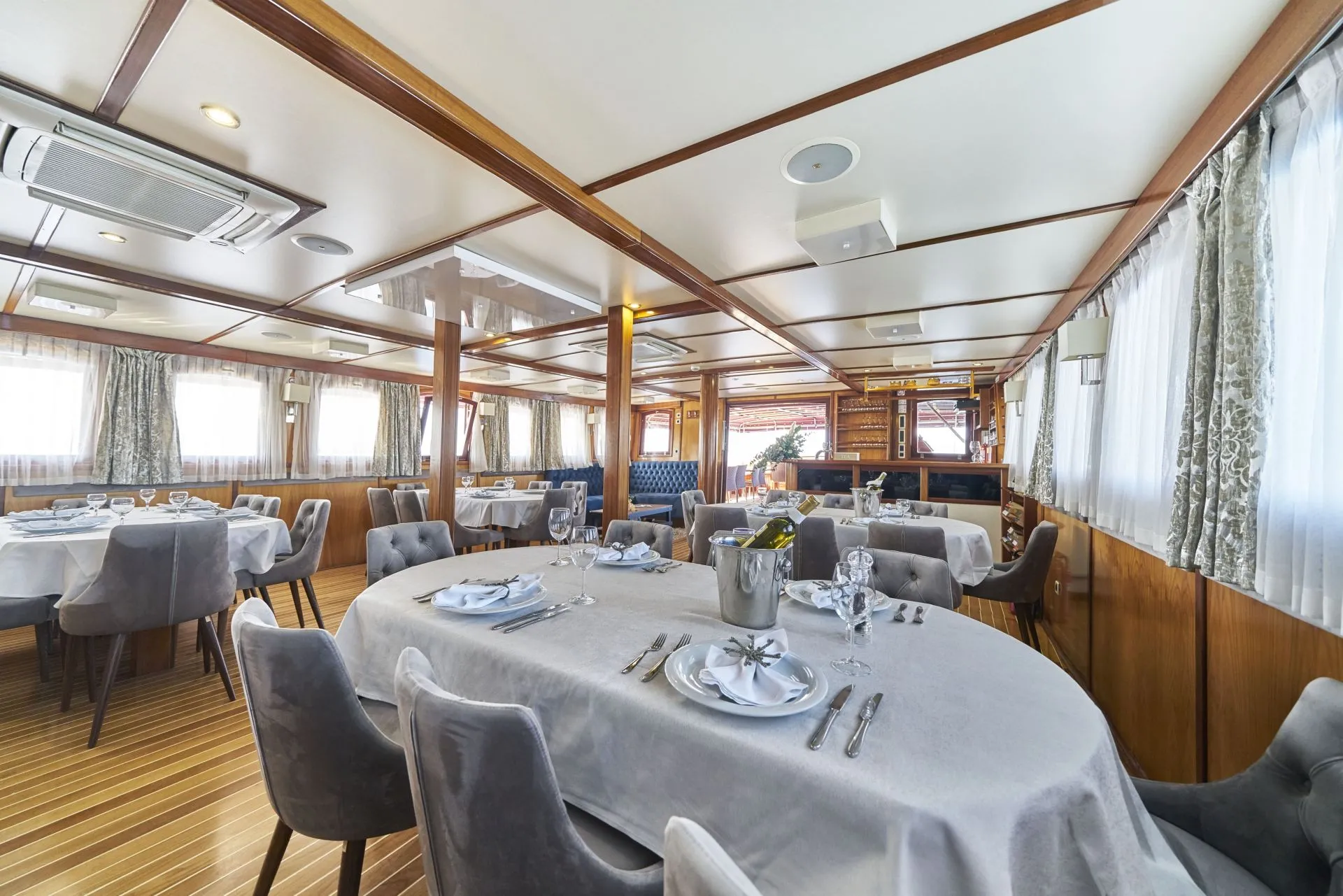 A spacious, elegant dining room with round tables covered in white cloths, gray upholstered chairs, set with glassware and cutlery. Large windows with curtains let in natural light, and wood paneling decorates the room.