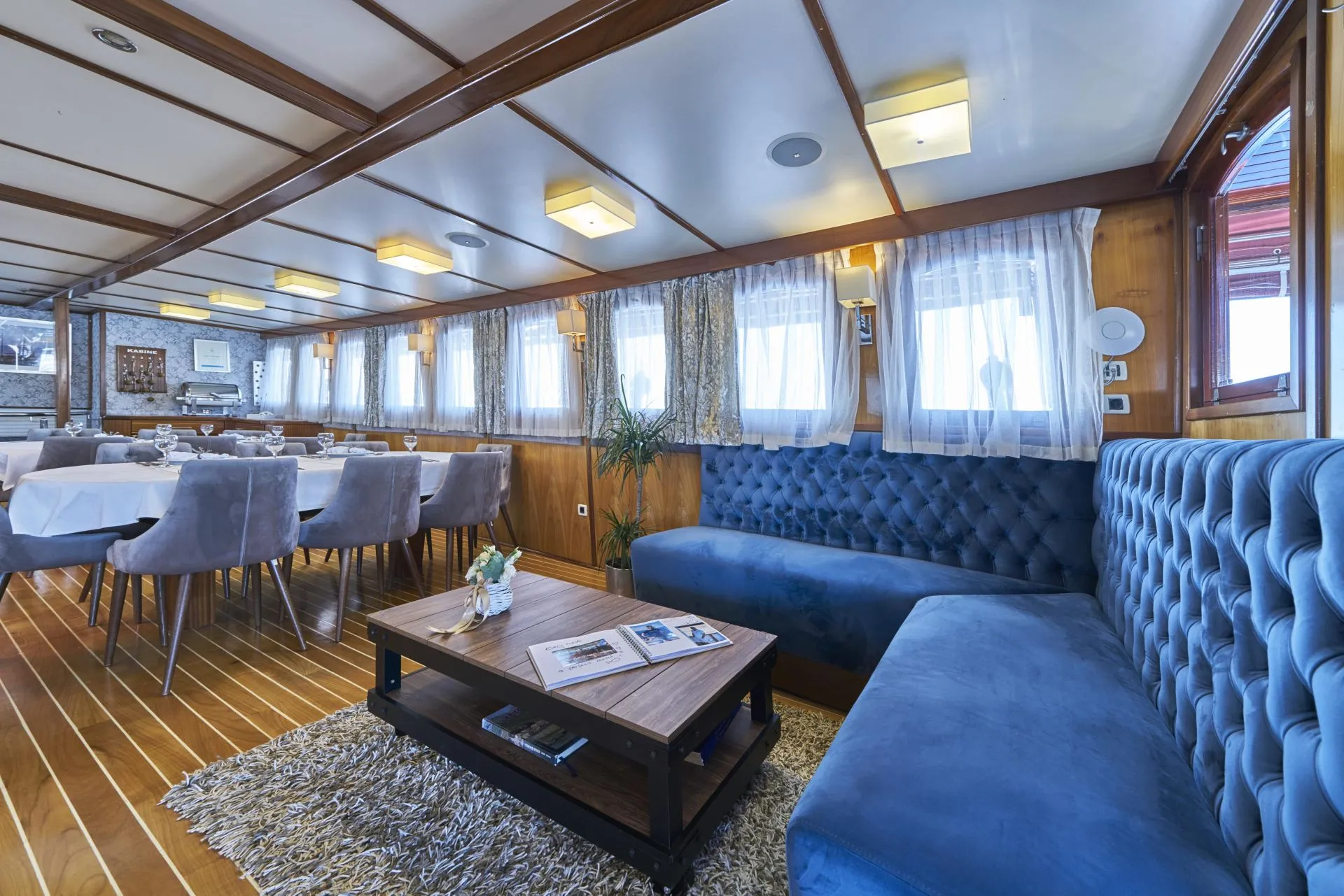 A spacious, elegantly decorated dining lounge on a boat features blue tufted seating, a wooden table with magazines, grey chairs around white-clothed tables, wood-paneled walls, and large windows with sheer curtains.