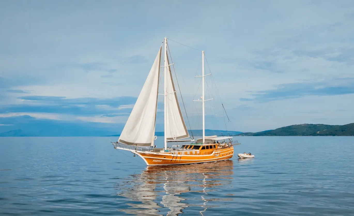 A wooden sailboat with white sails floats on calm, blue water under a cloudy sky, with green hills visible in the distance. The boats reflection is visible on the smooth surface of the sea.