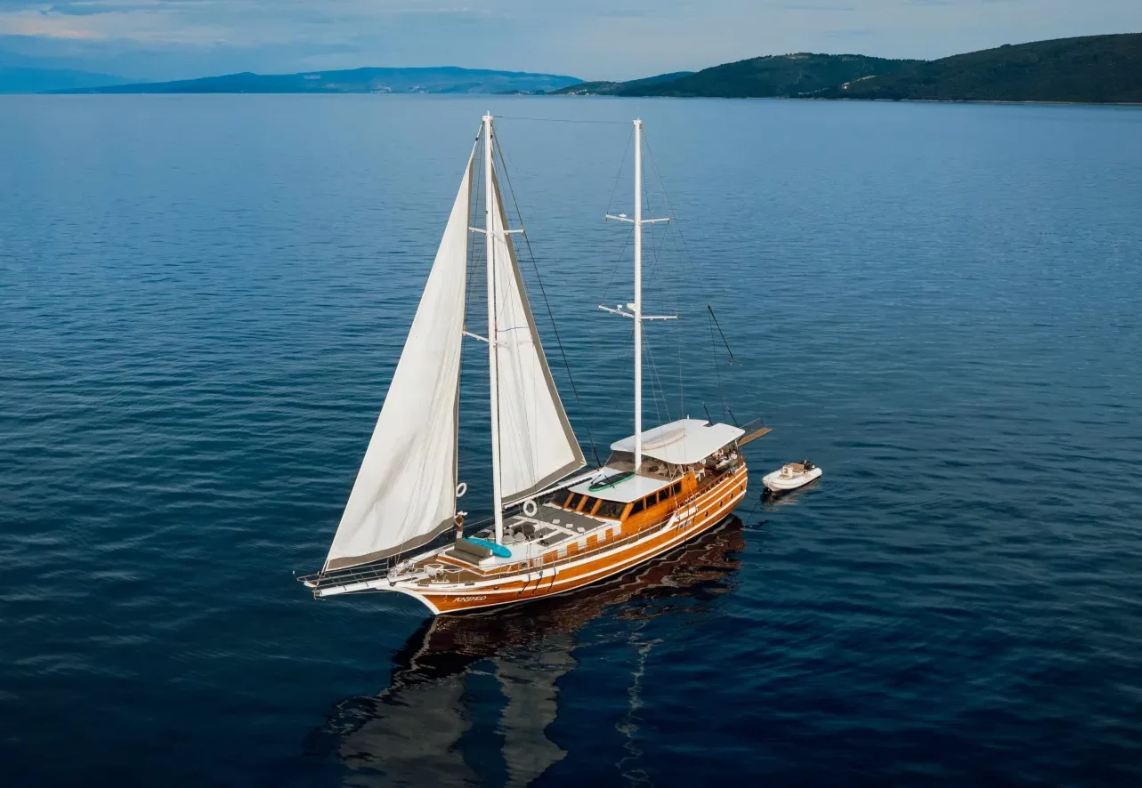 A wooden sailboat with two masts and white sails glides across calm, deep blue water. The boat is surrounded by open sea, with distant hills and a cloudy sky on the horizon.