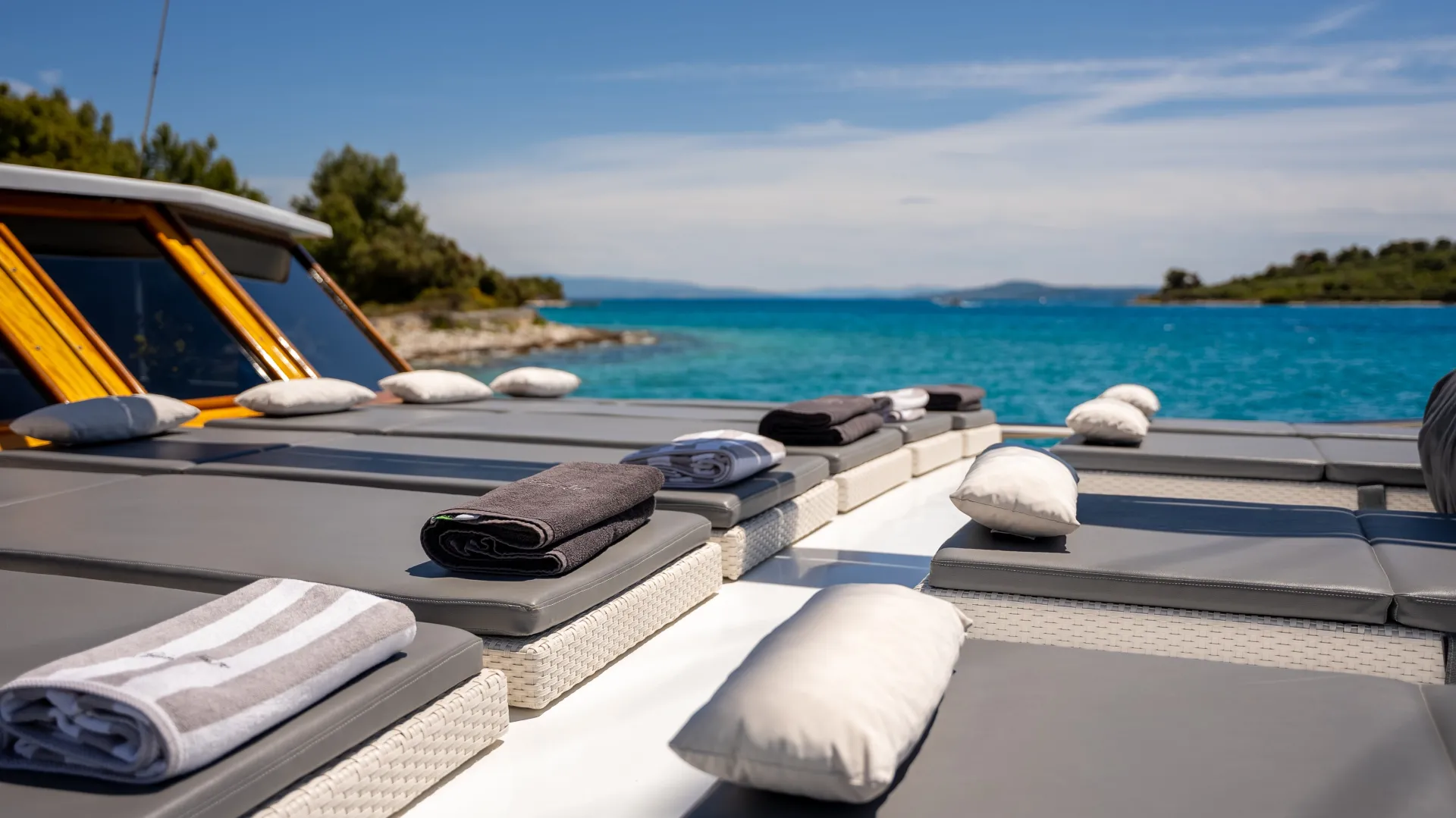 Sun loungers with folded towels and white pillows are arranged on the deck of a boat, overlooking calm turquoise water and a distant shoreline under a sunny, partly cloudy sky.