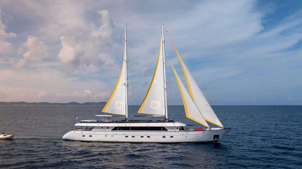 A large white luxury yacht with two tall masts and white sails edged in yellow sails across calm blue water under a partly cloudy sky. A small boat travels nearby in the background.