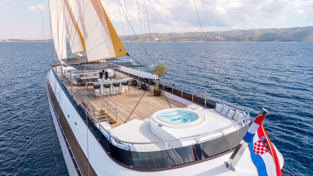 Luxury yacht with sun deck, hot tub, and seating area sailing on calm blue water near a mountainous coastline, flying the Croatian flag at the stern under a partly cloudy sky.