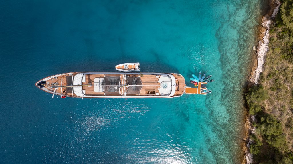 A large yacht is anchored in clear blue water near a rocky, tree-lined shore. Several kayaks and a small boat are next to the yacht, and the scene is viewed from above.