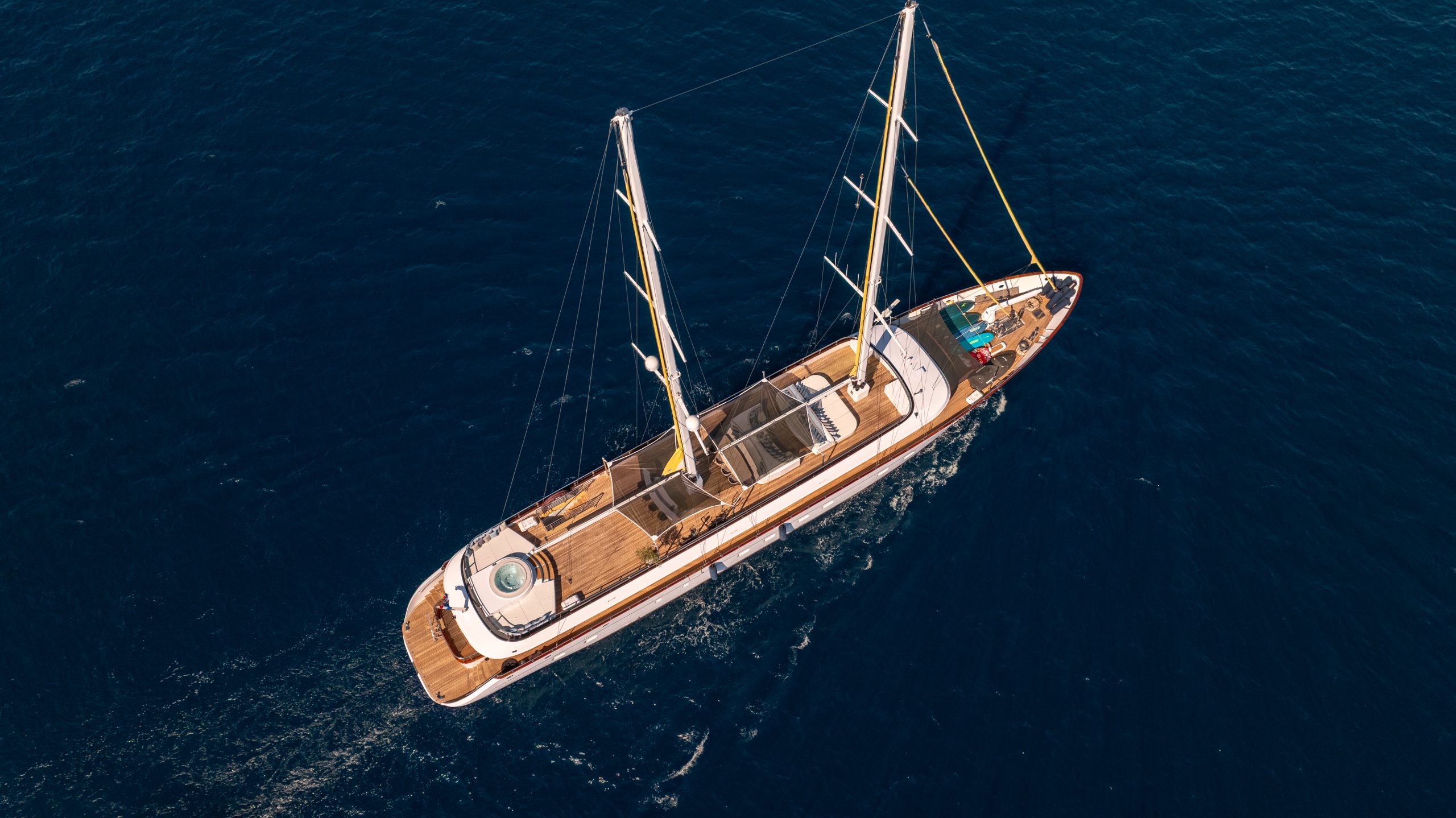Aerial view of a luxurious sailboat with two masts cruising on deep blue water, featuring wooden decks, lounging areas, and a small pool or hot tub at the back.