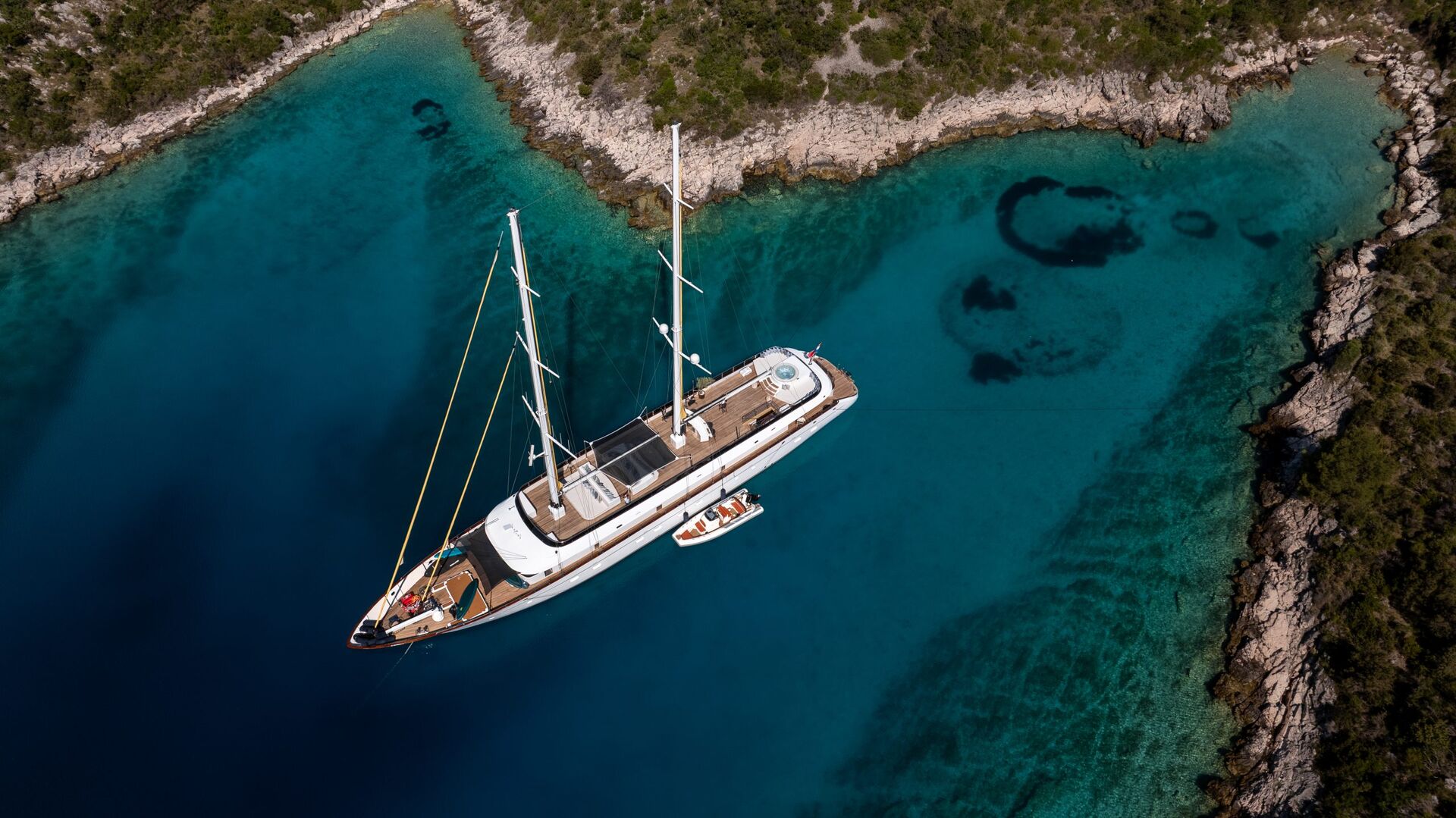 Aerial view of a large sailboat anchored in a clear turquoise bay surrounded by rocky shoreline and green vegetation, with a small boat floating next to it.