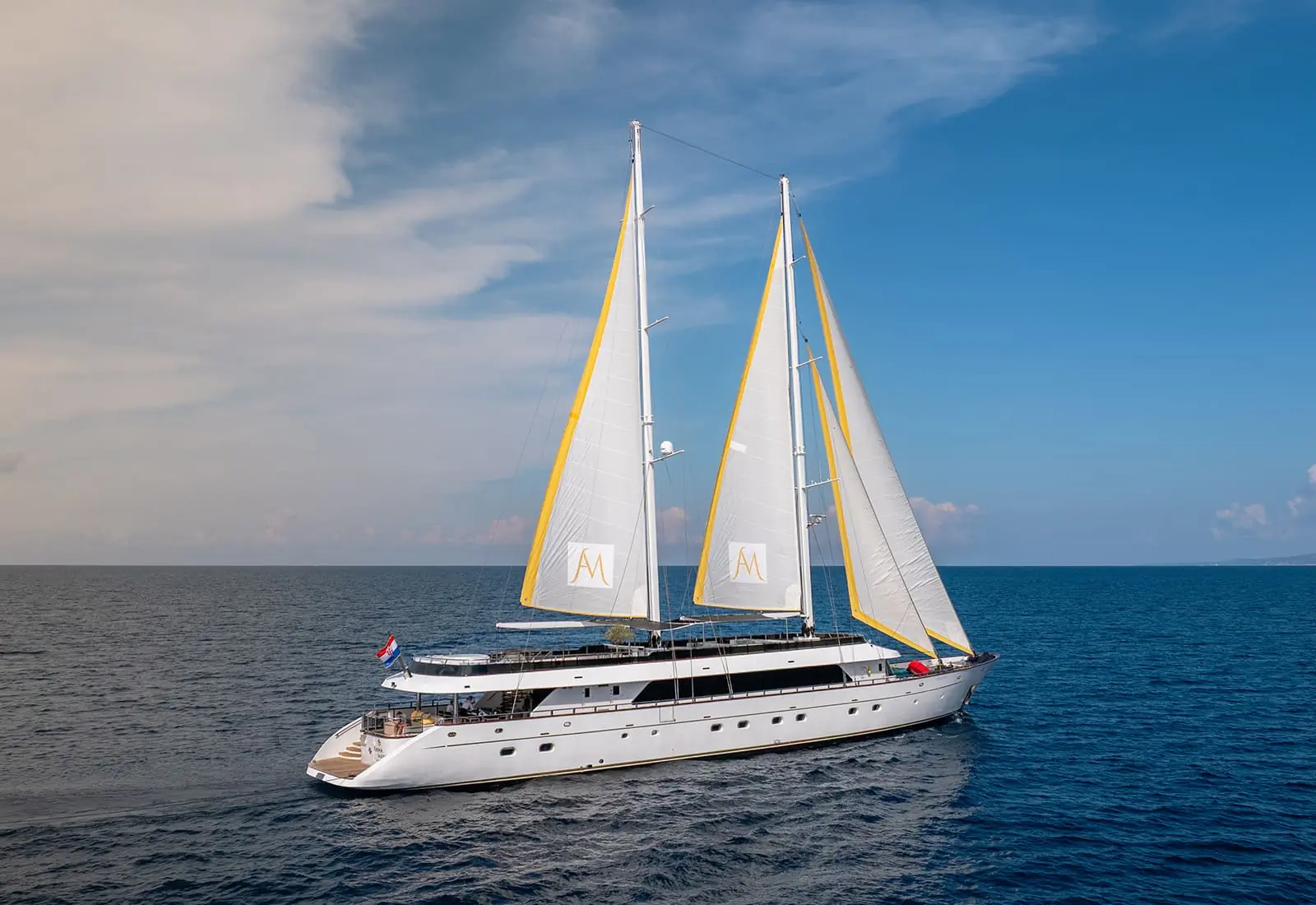 A large white sailing yacht with two yellow-trimmed masts sails on calm blue ocean water under a partly cloudy sky.