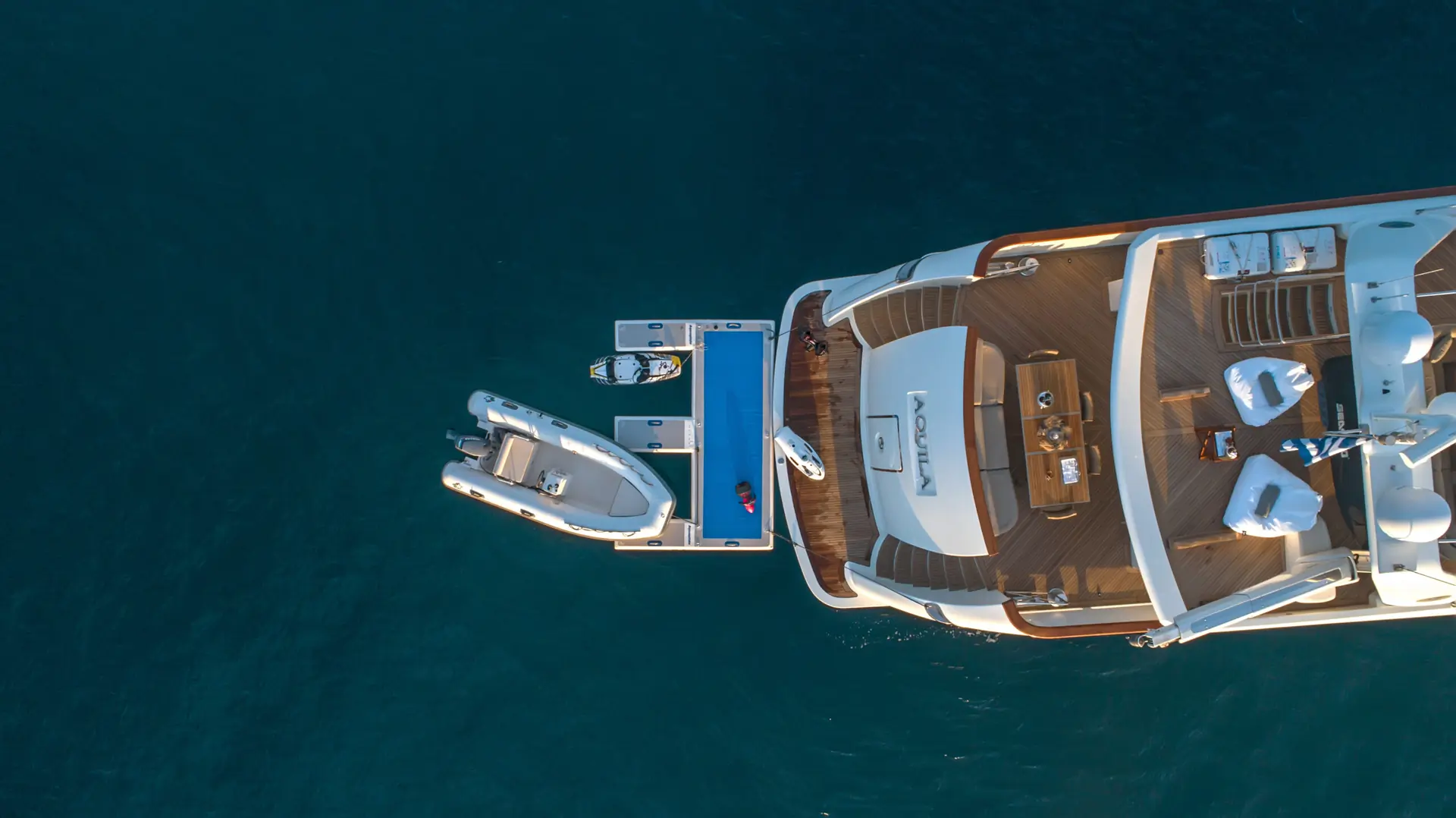 Aerial view of a luxury yacht with a smaller boat and jet ski docked alongside, floating on calm blue water. Lounge chairs and tables are set up on the yachts deck.