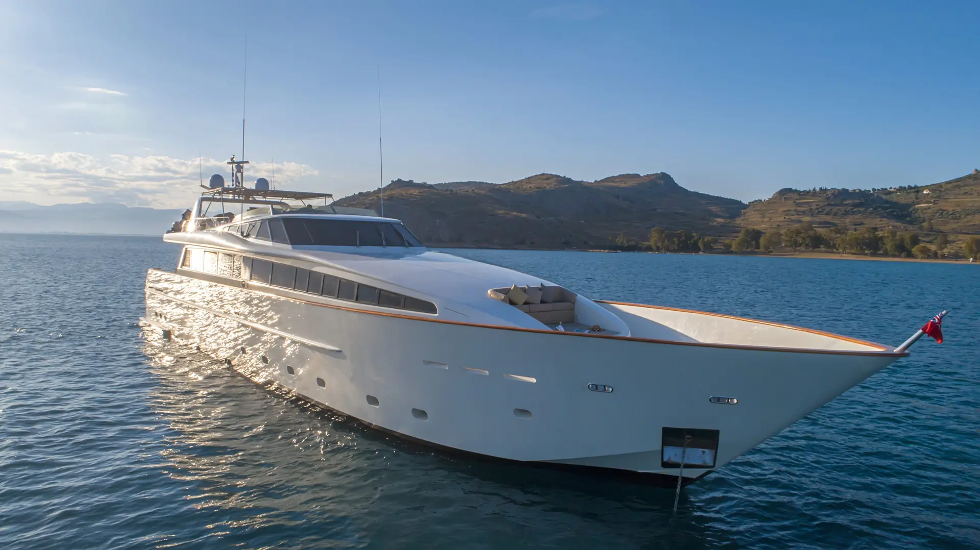 A sleek white yacht floats on calm blue water near a coastline with hills under a clear sky, reflecting sunlight on its surface.