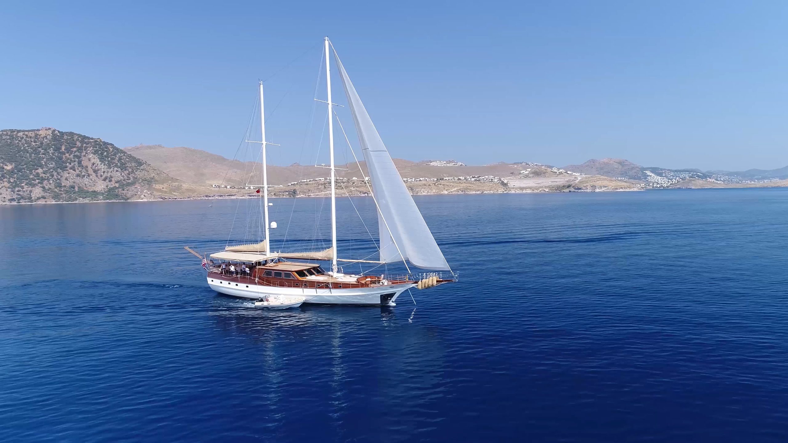 ARABELLA A large sailboat with white sails glides on calm, deep blue water near a coastline with hills and scattered buildings under a clear blue sky.