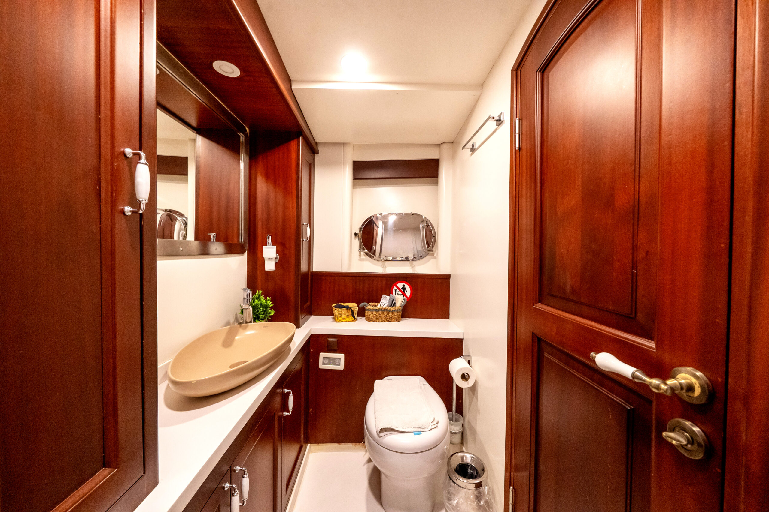 VIRTUOSO A small bathroom with wooden cabinets and doors, a beige sink, a toilet, a countertop with toiletries, a round mirror, and a small window letting in natural light.