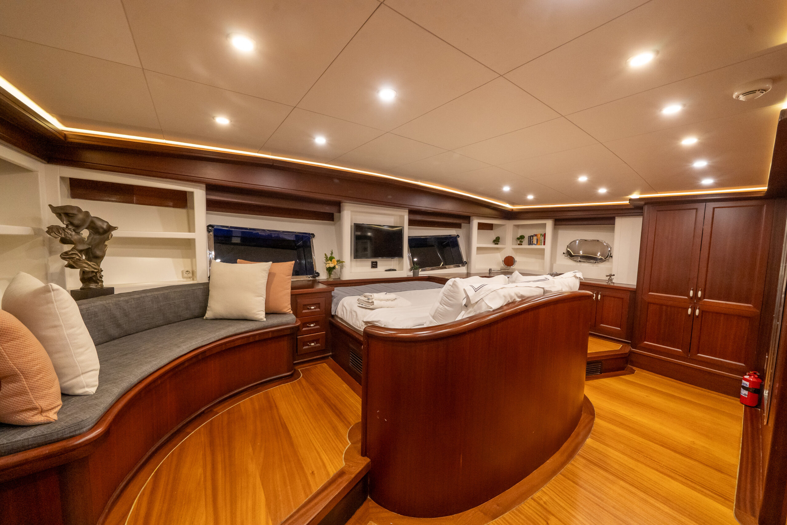 VIRTUOSO Luxurious yacht cabin with polished wood floors and trim, a large bed with white linens, a curved built-in sofa with cushions, shelves with decor items, and warm recessed lighting on the ceiling.